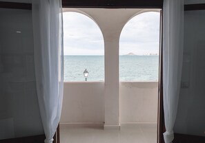 A room with a view of the sea through a balcony with white curtains.