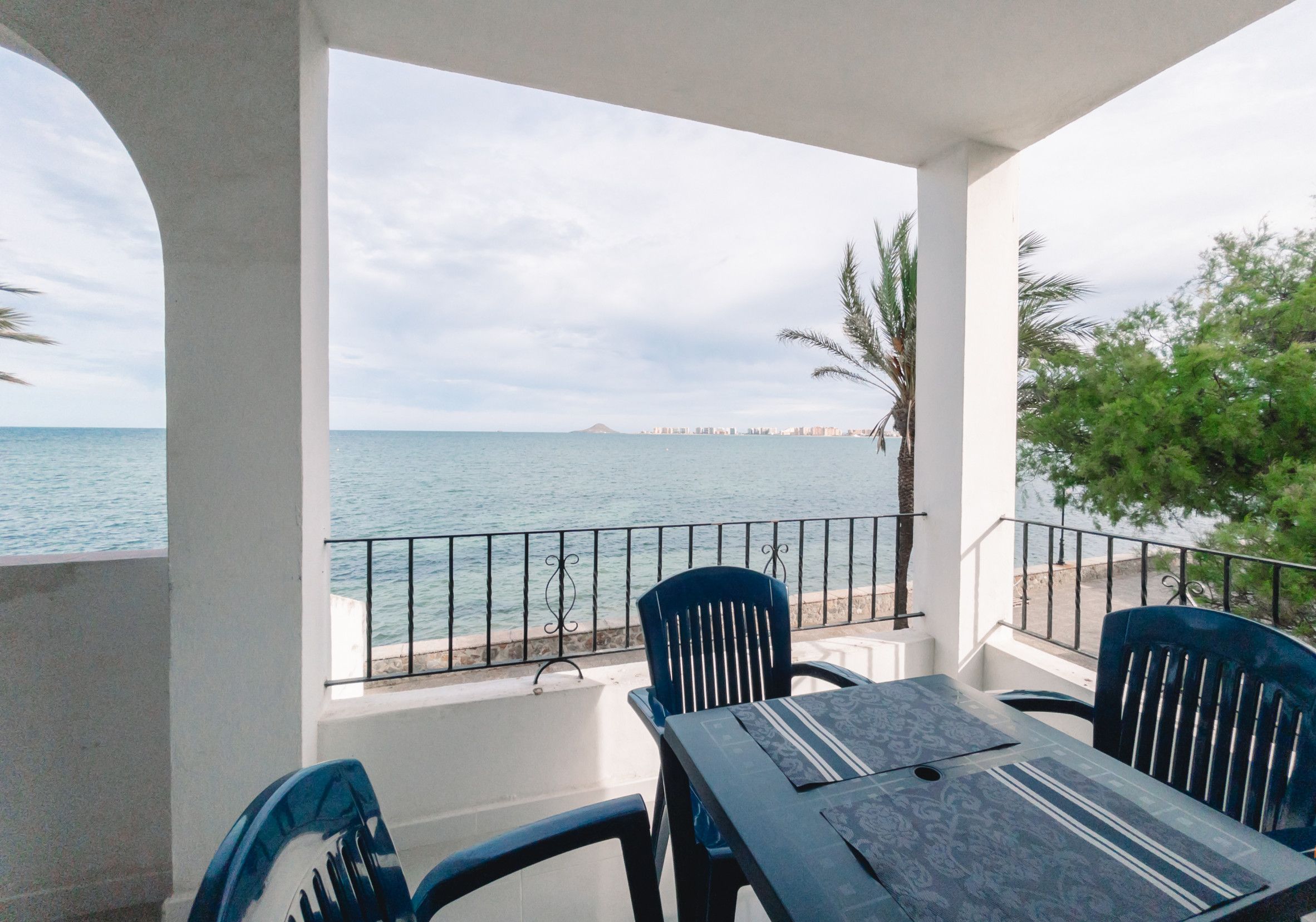 Ein Balkon mit Tisch und Stühlen, der auf eine Küstenlandschaft mit Geländer und Palmen blickt.