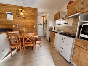 A compact kitchen with wooden cabinets, a dining table with chairs, a hanging light fixture, and a refrigerator.