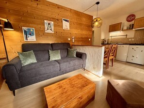 Una sala de estar moderna con un sofá gris, una mesa de centro de madera y una cocina con armarios blancos y una lámpara colgante.