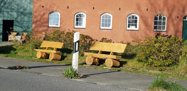 Un bâtiment en briques rouges, doté de fenêtres en arc de cercle et d’une porte verte. On y trouve deux bancs en bois sur lesquels sont posées des bûches, ainsi qu’une petite plante devant les bancs.