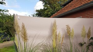 A wicker chair with a star-patterned pillow, a metal watering can, and tall grasses against a wall. - Apartment "Blied" f. Max. 2 pers + toddler, san. Gulfhof, gr. garden (Manslagt)