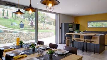 Een moderne keuken met een centraal eiland, een eethoek met een tafel voor vier personen, en grote ramen met uitzicht op de omgeving.