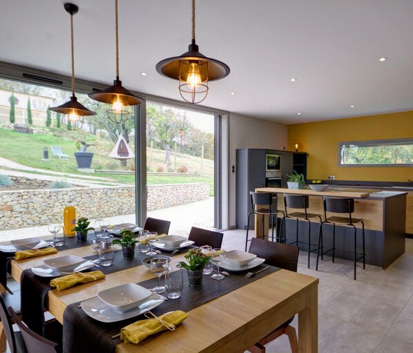 Une cuisine moderne dotée d’un îlot central, d’un coin repas avec une table pour quatre personnes et de grandes fenêtres offrant une vue sur l’extérieur.