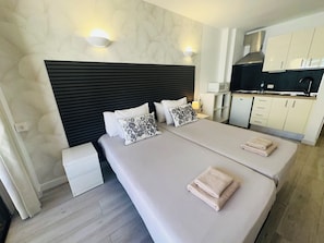 A modern hotel room with a large bed, a bedside table, a built-in kitchen area, and a wall-mounted light fixture.