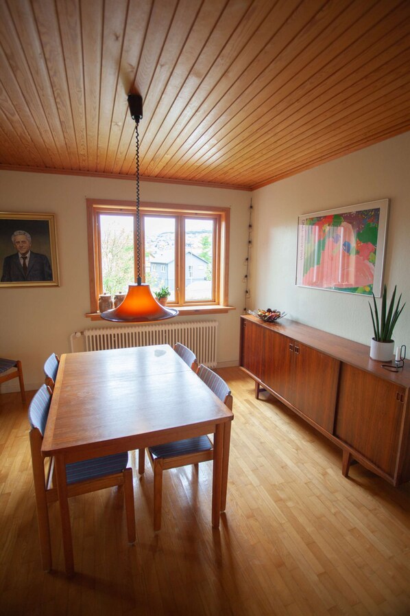A dining area with a wooden table, chairs, a hanging orange pendant light, a window with a view, a radiator, a cabinet, and a framed portrait on the wall.