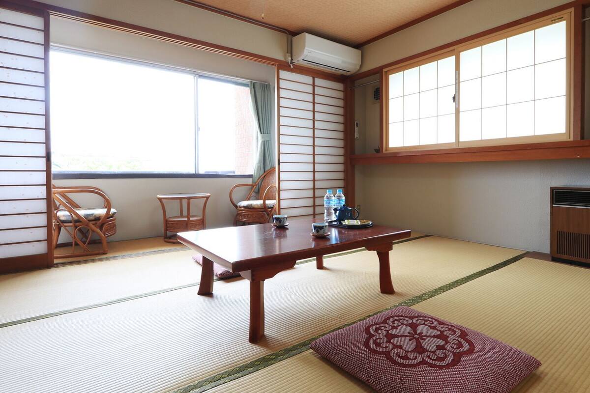 A traditional Japanese room with tatami flooring, a low table, two chairs, a futon, and a radiator.