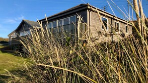 A rustic wooden house with a sloped roof, surrounded by tall grass and vegetation. - E46 Dunroven, Riviere Towans (Hayle)
