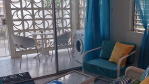 A room with a sofa, a chair, a table, a washing machine, and a window with curtains. - Beachside Apartment #2 - steps from the beach! (Bridgetown)