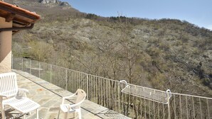 Un balcón con suelo de piedra, sillas blancas y barandilla de metal con vistas a un paisaje montañoso.