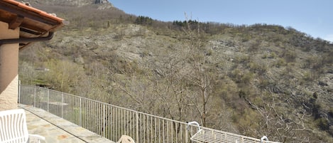 A balcony with stone flooring, white chairs, and a metal railing overlooking a mountainous landscape.