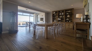 Un comedor amplio con una mesa de madera y sillas de plástico transparente, un aparador de madera con objetos decorativos, y vistas a través de un ventanal grande.