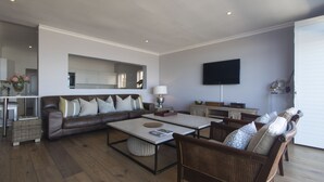 Una sala de estar moderna con un sofá de cuero marrón, una mesa de centro, sillas de mimbre, un televisor de pantalla plana montado en la pared y una zona de cocina al fondo.