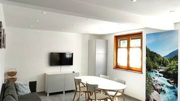 A modern living room with a white sofa, a dining table with chairs, a flat-screen TV mounted on the wall, and a large window with a view of a river and mountains.
