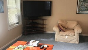 A living room with a flat-screen TV, a beige armchair, a brown couch, and a coffee table with remote controls and magazines. - Spacious Condo on Sunday River Access Road (Bethal)