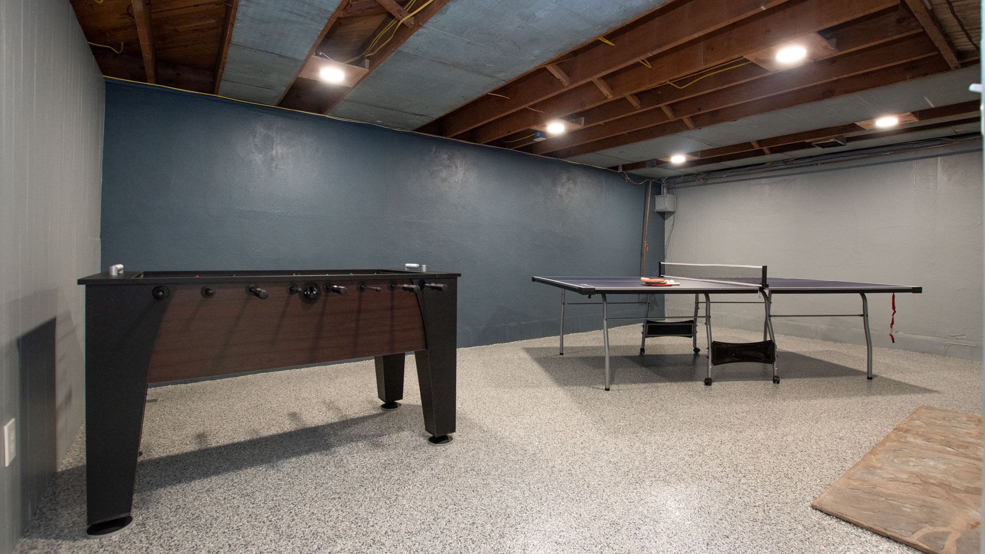 Una habitación con mesa de billar y mesa de ping pong, vigas de madera en el techo y suelo cubierto de alfombra gris.