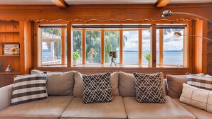 A cozy wooden cabin with a large window overlooking a lake, a cushioned sofa with patterned pillows, and a small table with a lamp. - Long View on the Cove: Views! Views! Views! (Seneca Falls)