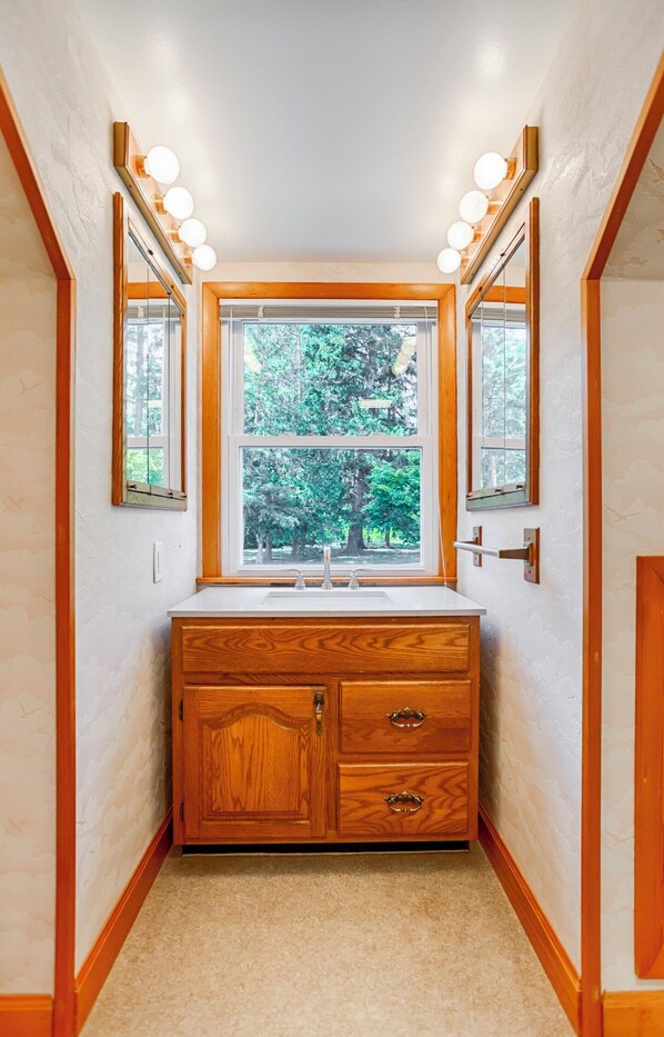 Een badkamer met een houten wastafel, een raam met uitzicht op bomen en twee spiegels met verlichting.