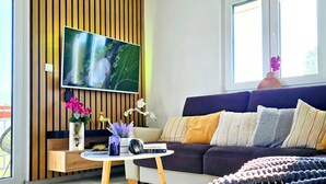 A modern living room with a flat-screen TV mounted on a wooden panel wall, a sofa with multiple cushions, a coffee table with a speaker, and a large window with a view of greenery. - Ferienhaus Biograci near Mostar – Modern Comfort & Nature (Siroki Brijeg)