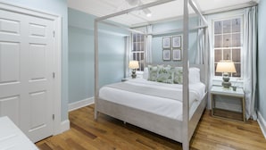 Una cama con dosel y cuatro columnas, una mesita de noche con lámpara, una ventana con cortinas y una puerta con marco blanco.