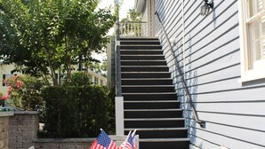 Une terrasse en bois ornée de deux drapeaux américains, un mur de pierres et un escalier menant à un bâtiment dont le revêtement extérieur est constitué de planches disposées horizontalement.