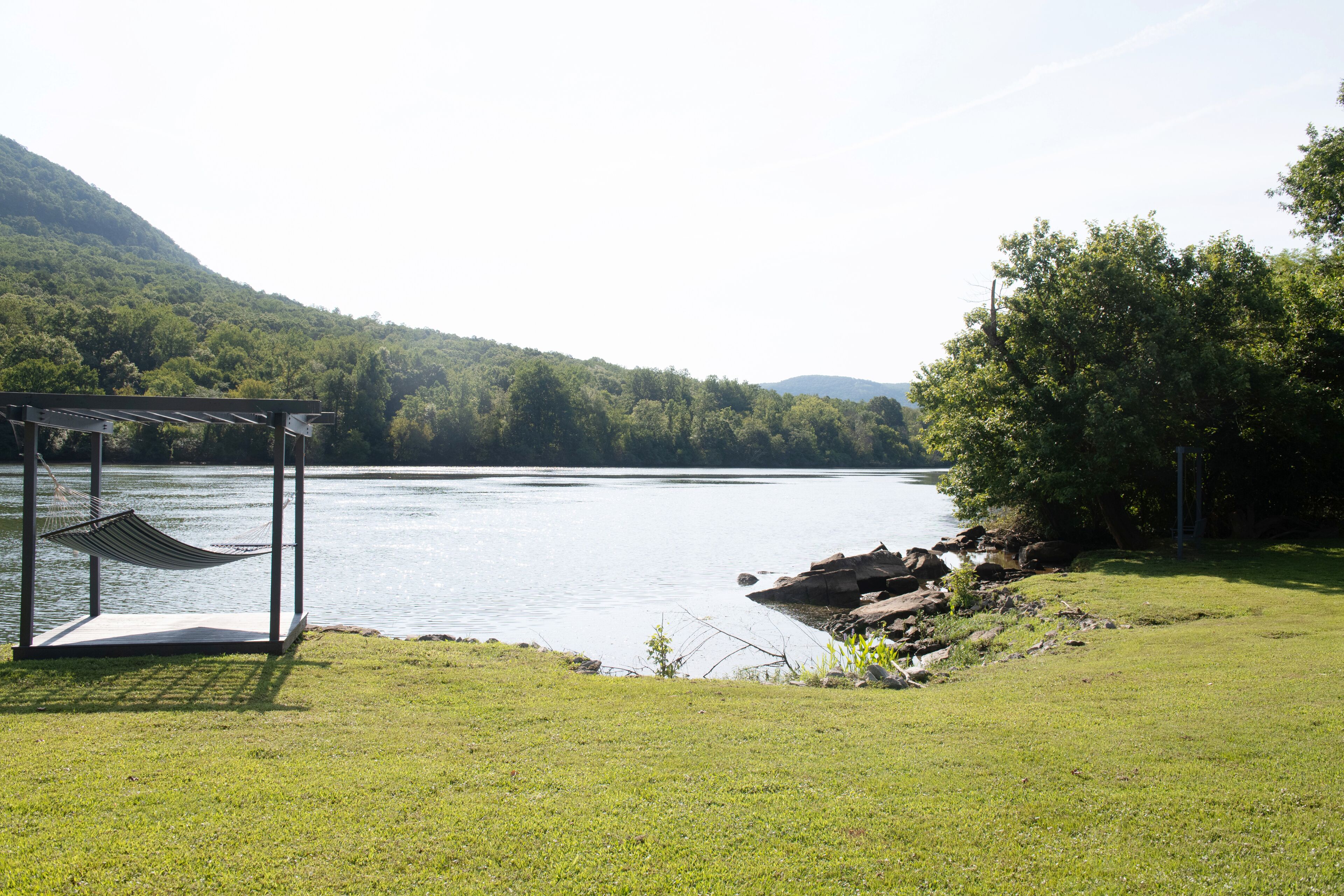 A hammock under a gazebo by a lake with a forested hillside in the background.