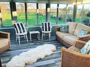 A sunlit room with wicker furniture, a white furry rug, and a view of a green field through large windows. - 4 Bed Beach Bungalow with Dog-friendly Garden & Hot Tub on South Beach, Heacham (Heacham)