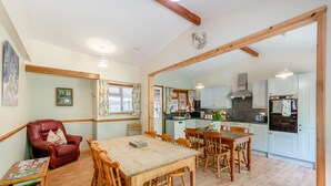 Un coin repas avec une table et des chaises en bois, un fauteuil en cuir rouge, une petite table d’appoint avec des magazines, et une cuisine en arrière-plan.