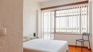 A bedroom with a bed, a chair, and a window with blinds. - House on the beach in Sesimbra (Sesimbra)