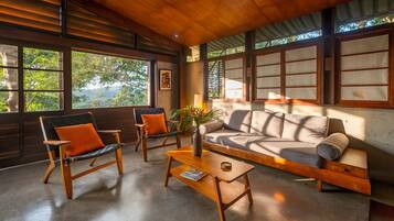 A modern living room with a sofa, two chairs, a coffee table, and large windows offering a view of trees and a body of water.
