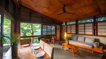 A wooden-floored living area with a sofa, chairs, a dining table set for two, and a ceiling fan.
