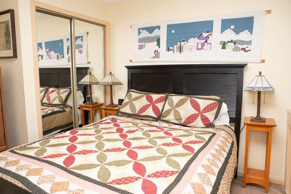 A bedroom with a bed, a nightstand, a lamp, a mirror, and a wall with a quilted tapestry.