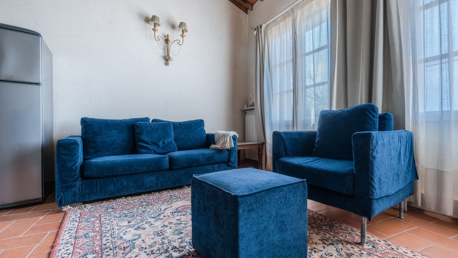 Una sala de estar con un sofá azul, un sillón a juego y un pequeño reposapiés. Hay una alfombra estampada en el suelo, un radiador y una ventana con cortinas.