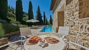 Escena junto a la piscina con un muro de piedra, una mesa con frutas y bebidas, y sillas de descanso.