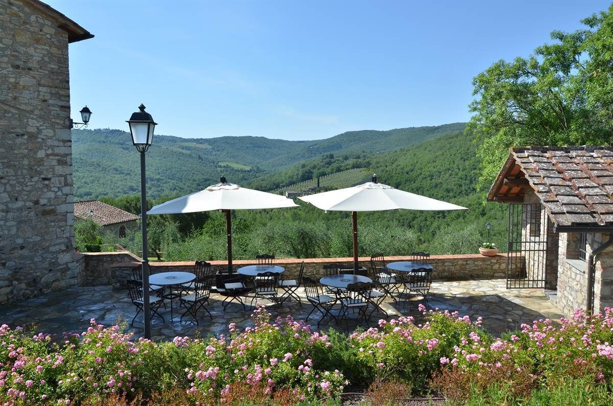 An outdoor patio with stone flooring, round tables, and chairs, surrounded by blooming pink flowers.
