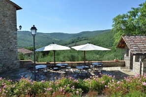 An outdoor patio with stone flooring, tables, and chairs, surrounded by blooming flowers and a view of green hills.