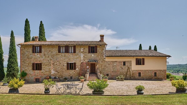 Une maison en pierre avec un toit en tuiles, un bâtiment séparé à la façade jaune, et un jardin bien entretenu agrémenté de plantes en pot et de mobilier d’extérieur.