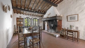 A rustic kitchen with a large wooden table, chairs, a fireplace, and a view of greenery through the windows. - Villa San Michele, Gaiole in Chianti, Siena and Chianti (Gaiole in Chianti)