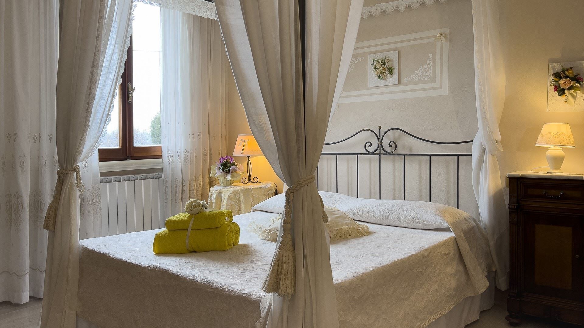Un dormitorio con cama con dosel, una mesita de noche con lámpara y una ventana con cortinas.