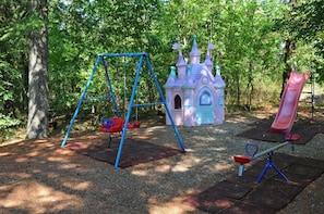 Explore the playground area with a swing set, castle play structure, and seesaw set amidst shaded trees.