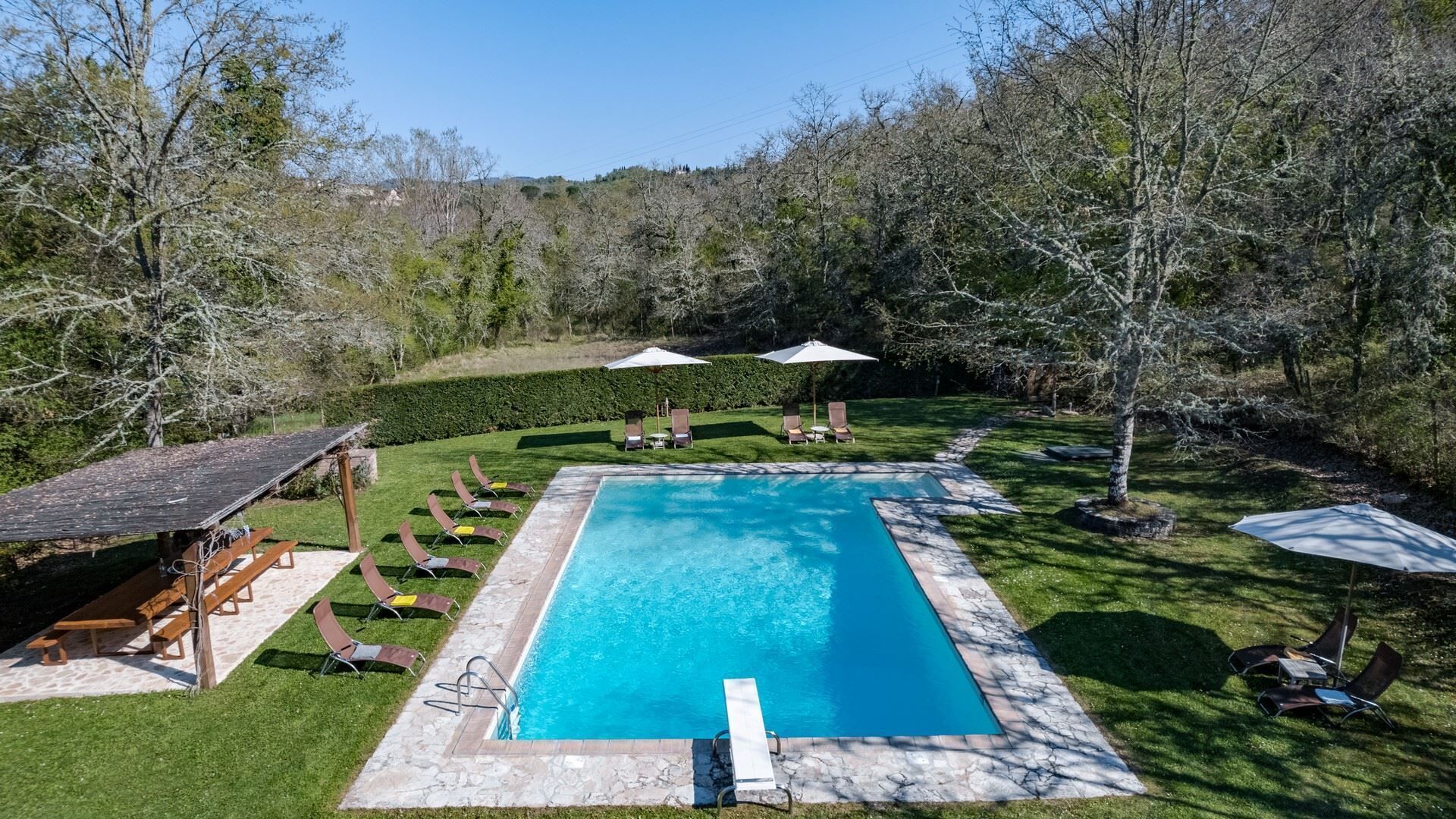 A rectangular swimming pool with a stone border, surrounded by a grassy area with lounge chairs and umbrellas.