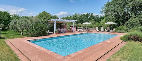 Una piscina rectangular con solárium de baldosas, rodeada de césped y árboles.