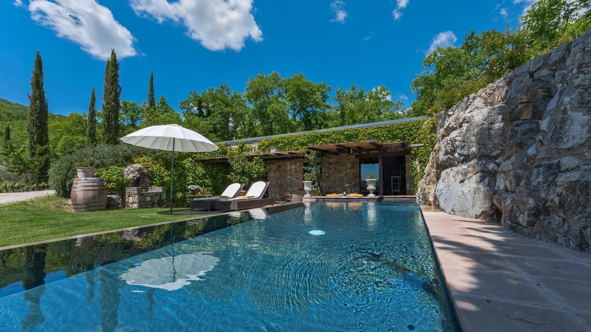 A clear swimming pool with lounge chairs and a pergola, surrounded by greenery and stone structures.