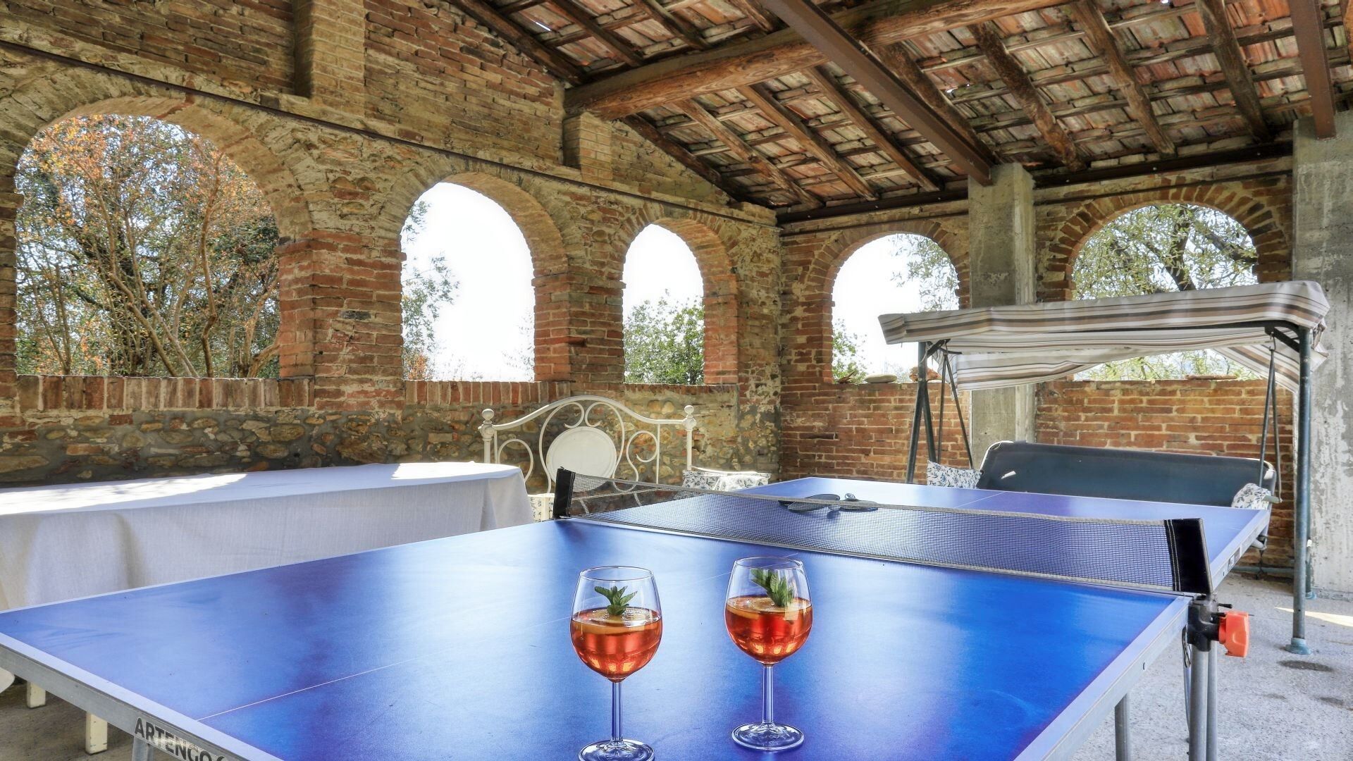 A table tennis table with two glasses of red beverage, a metal chair, and a canopy.