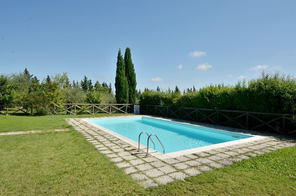 A rectangular swimming pool with a tiled deck, surrounded by a wooden fence and lush greenery.