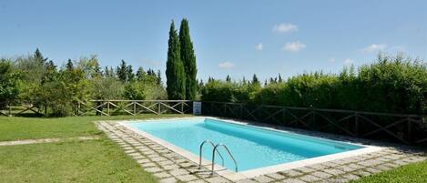 A rectangular swimming pool with a tiled deck, surrounded by a wooden fence and lush greenery.
