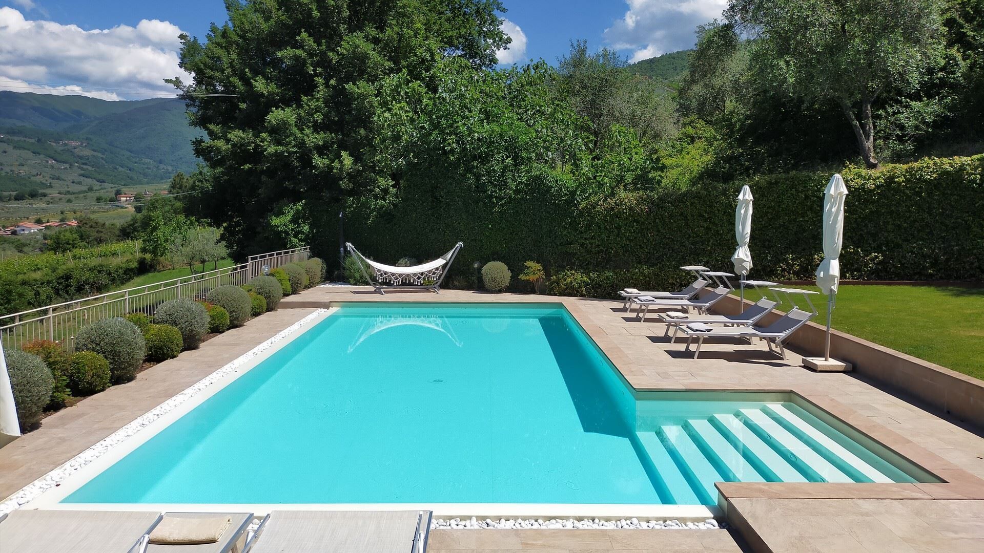A rectangular swimming pool with a hammock and lounge chairs, surrounded by well-manicured bushes and trees, with a view of distant hills.