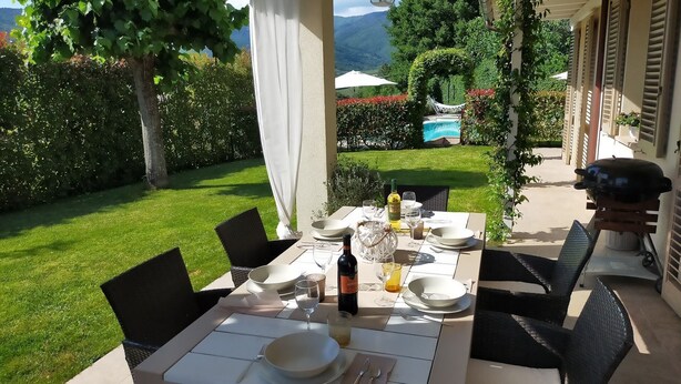 Un área de comedor al aire libre con una mesa preparada para cuatro personas, rodeada de sillas. Se ve una parrilla y una piscina al fondo.
