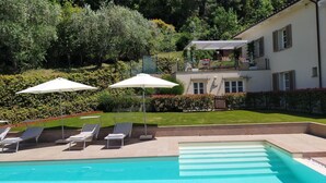 Una piscina con sillas de descanso y sombrillas, rodeada por un jardín cuidado y un edificio de dos pisos.