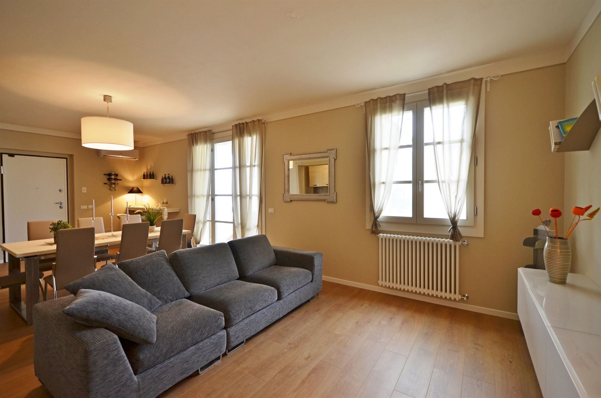 Un salon avec un canapé gris, un plancher en bois, un coin repas avec des chaises et une table, un radiateur et une fenêtre avec des rideaux.